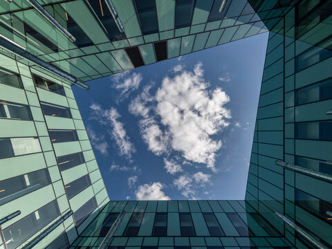 Low-angle view looking up through a rectangular courtyard formed by glass and metal office building facades, with exposed pipes along the walls. Clouds and blue sky fill the open frame at the center.