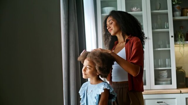 Mother combing curly afro hair daughter home kitchen. Morning grooming routine.