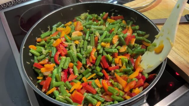 Close-up view of fresh green beans, sliced red and orange bell peppers, carrots, and onions being stirred with a wooden spatula in a frying pan on an induction stove.   