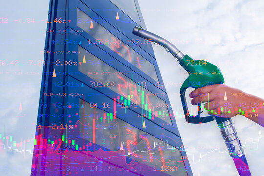 Hand holding green fuel nozzle near gas station sign board with stock market trading graph double exposure. Energy crisis, fuel price inflation, commodity market and stock trading concept.