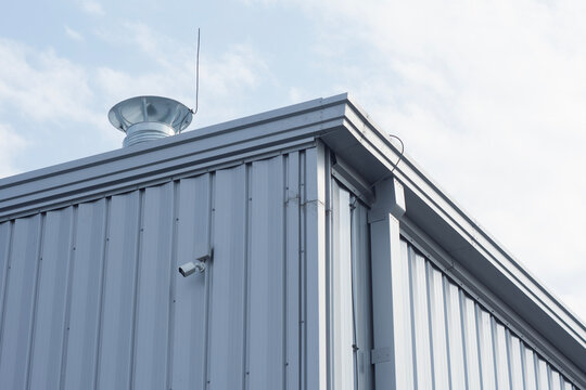 Modern industrial building exterior with corrugated metal cladding, wall-mounted security camera and rooftop ventilation turbine. Warehouse architecture, commercial property and CCTV.