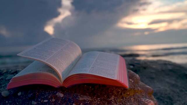 Open Bible Book with Fluttering Pages on Rocky Beach at Sunset