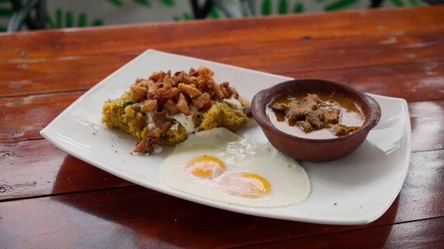 Green plantain tigrillo with pork cracklings, beef stew, and two fried eggs. Traditional Ecuadorian breakfast.