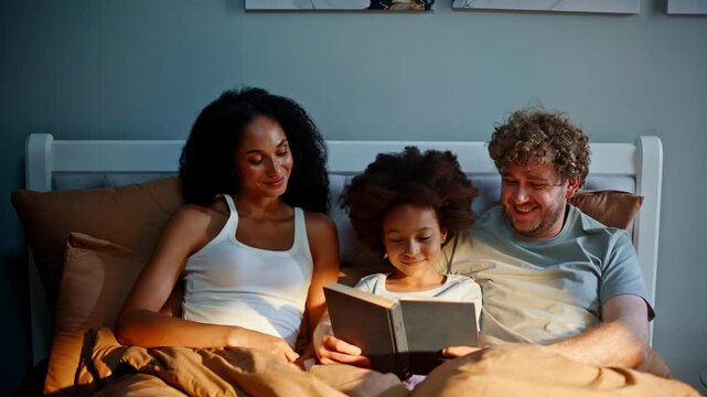 Multiracial family reading book bed night. Parents daughter bedtime story cozy.