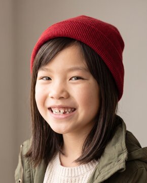 Playful Asian girl in red knit beanie and olive parka with wide grin, neutral studio backdrop