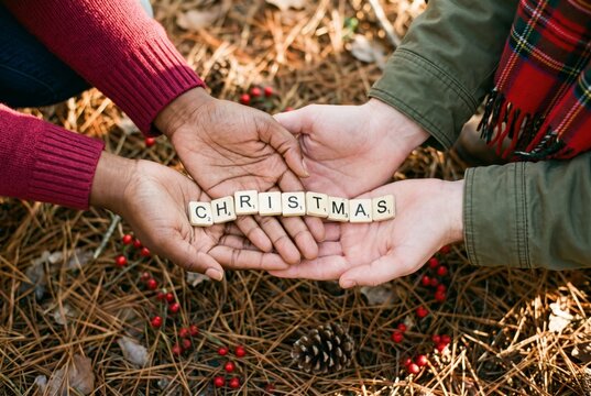 Diverse hands joined holding CHRISTMAS letter tiles over pine needles and berries, symbol of unity