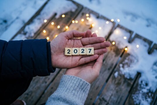 Cupped hands cradling 2027 number tiles over snowy deck with fairy lights, symbol of quiet hope