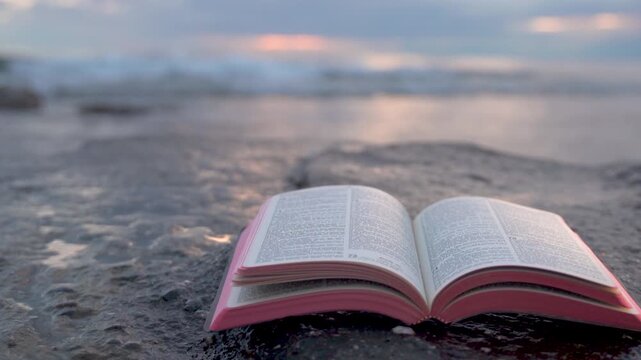 Open Bible Book with Fluttering Pages on Rocky Beach at Sunset