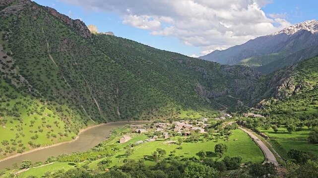 Mountain village nestled in a lush sunlit valley beside a winding river, with road, green meadows, yellow wildflowers, tree-covered mountains, fluffy clouds and distant snow-dusted peaks.
📍Zom