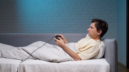 Young man relaxing in bed and playing video games using a wired controller, enjoying leisure time and entertainment in a modern bedroom with blue light © Aliaksandr