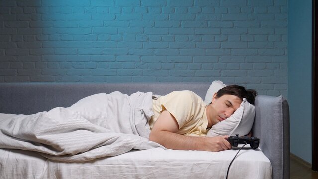 Young man sleeping deeply on a bed or sofa holding a video game controller, depicting exhaustion and burnout from gaming addiction or spending too much time playing