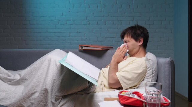 Sick man working from home on a sofa, blowing his nose amid papers, medicine and water, wrapped in a blanket while battling flu like symptoms and fatigue during remote work