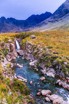 Waterfall cascades down rocky slopes into clear blue stream. Mountains rise sharply against soft purple sky. Lush green grass surrounds winding river. Stones line riverbed, reflecting sunlight. No peo