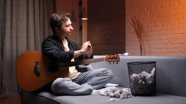 Young man sitting on a sofa with an acoustic guitar, expressing frustration and tossing crumpled papers towards a waste bin, symbolizing a creative block and songwriting struggle