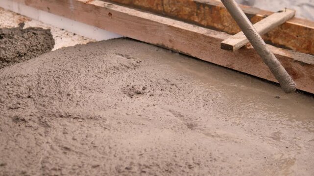 Fresh wet concrete spreads inside wooden formwork while diagonal screed tool levels surface at construction site, with rough slurry texture, damp sheen, weathered timber, and warm natural light.