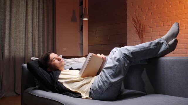 Young adult man lying comfortably on a modern gray sofa at home, taking a break from reading and looking contemplatively upwards during a quiet evening