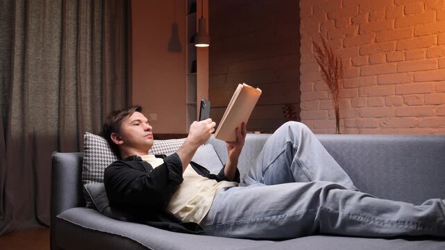 Young man asleep on a gray sofa in a dim living room, dressed casually after reading a book, resting peacefully during an evening nap in his modern apartment