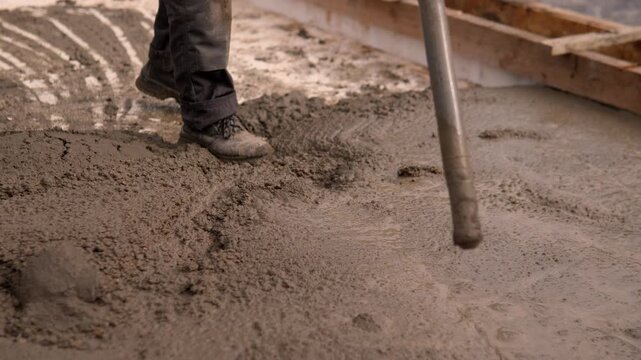 Construction worker boots and lower legs press through fresh concrete while long handle tool spreads thick slurry across unfinished foundation.