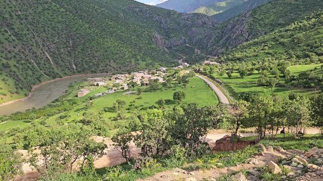 Mountain village nestled in a lush sunlit valley beside a winding river, with road, green meadows, yellow wildflowers, tree-covered mountains, fluffy clouds and distant snow-dusted peaks.
📍Zom