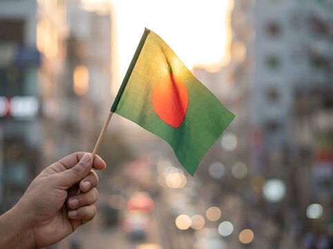 Hand holding a small bangladesh flag against a blurred urban background with golden backlight bangladesh background
