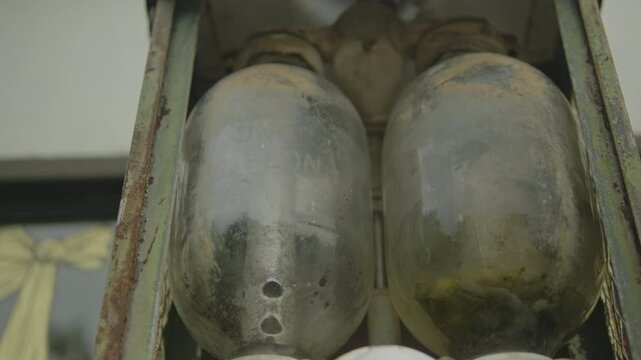 Low-angle close-up shows two dusty, weathered glass bulbs mounted in a rusted metal frame and cage, revealing corrosion, patina and surface grime.