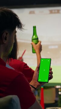 Vertical frame: Supporter checks live scores on a green screen smartphone betting app during a rooftop match projection, while friends enjoy the soccer game nearby.