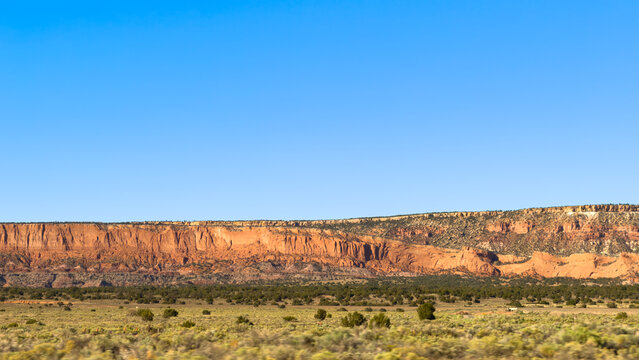 Sunlit red-orange sandstone escarpment above a juniper belt and sagebrush flats, motion-blurred foreground, crisp cloudless blue sky