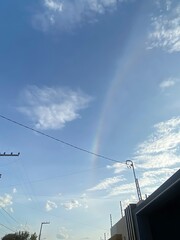 Fototapeta premium A faint rainbow arcs across a bright blue sky with scattered clouds and overhead wires