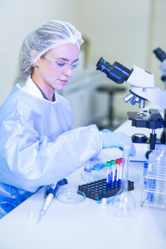 A woman lab technician in white laboratory clothing, safety glasses and blue gloves withdrawing hand from filled tube rack at a brightly lit laboratory workbench.