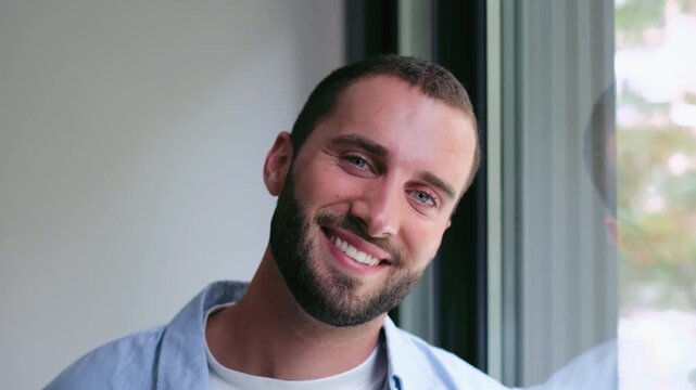 Young man leans by a window at home, looks outside thoughtfully, then turns and smiles at the camera. Casual indoor portrait captures a relaxed, cheerful lifestyle with soft natural window light.