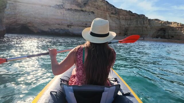 Woman paddles an inflatable kayak across calm turquoise water toward a sandy beach, passing rugged cliffs and caves. Handheld tracking shot of a summer travel and outdoor adventure.