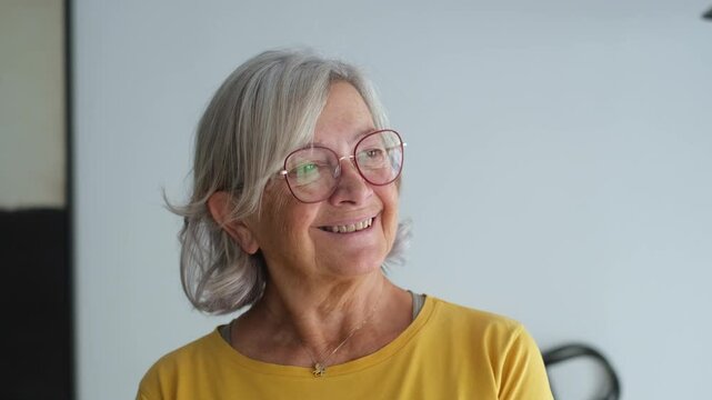 Senior woman with grey hair and glasses smiles at the camera, then turns her gaze aside indoors. Her cheerful expression conveys active aging, retirement, wellness and a positive senior lifestyle.