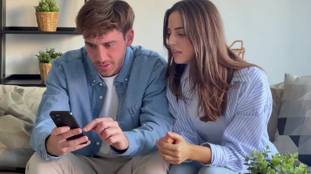 Young couple sits together on a sofa at home, discussing smartphone content. Man scrolls and points at the screen while woman leans close, talks and touches his arm during shared online browsing.