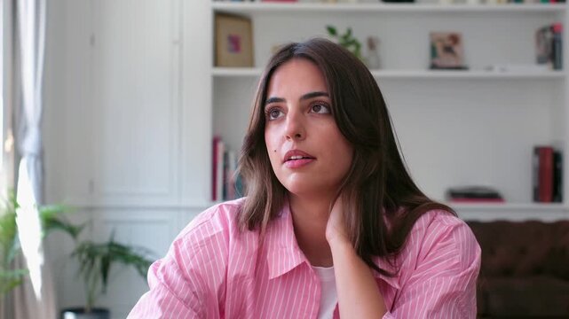 Bored young woman sighs, shifts her pose and rests her chin on her hand in a bright living room. Her tired, thoughtful gaze wanders around the room, conveying fatigue, boredom and low motivation.