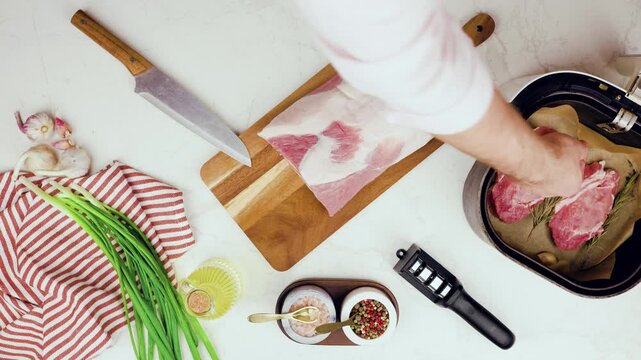 Chef preparing raw pork meat slices in air fryer with garlic and herbs. Top view of cooking pork chops with fresh seasonings, oil and spices on kitchen wooden board countertop now. 4k footage