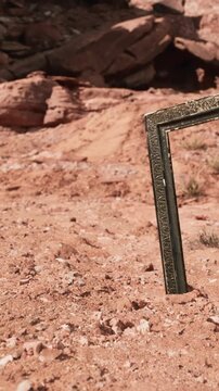 A metal frame stands in a desert area with red soil and sparse vegetation. The frame is tilted and partially buried. The location appears dry and rocky, suggesting a remote setting.
