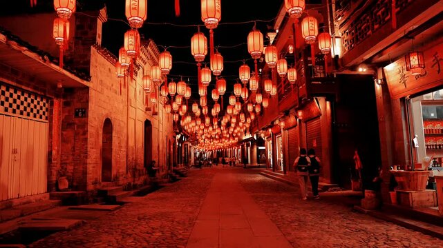 Red Chinese New Year lanterns hanging in rows over a cobblestone historic alleyway at night, with pedestrians walking beneath glowing traditional lanterns