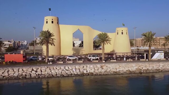 Aerial drone view of the historic Al Jazirah Al Hamra gate with Saudi Arabian flags, palm trees, and waterfront stone embankment under clear blue sky