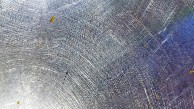 A close-up view of a scratched metal surface&mdash;possibly stainless steel or aluminum&mdash;bearing faint traces of yellow paint. The texture is rough, featuring a multitude of fine and deep scratches crisscros