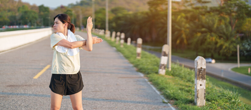 Asian woman with a towel around her neck cooling down and stretching after jogging in the park. Female runner relaxing after morning workout. Fitness, recovery and healthy lifestyle concept