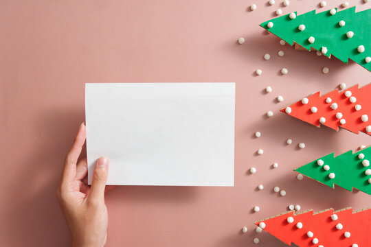 Cropped hand holding blank white card mockup on pink background with paper Christmas trees