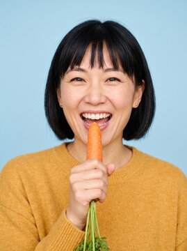 Cheerful East Asian woman in mustard sweater holding fresh carrot like a microphone with big excited smile on plain soft sky blue background isolated close up portrait for playful food concepts