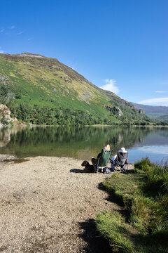 People relaxing in folding chairs on the gravel shore of Llyn Gwynant with a dog, enjoying views of the tranquil lake and surrounding mountains in Snowdonia National Park, North Wales.