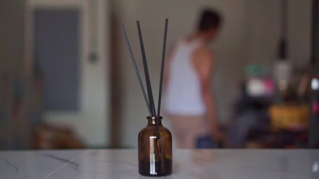 Reed diffuser sticks in an amber bottle at home with blur people activity on the background. Lifestyle footage ideal for aromatherapy, spa, relaxation, and home fragrance content.