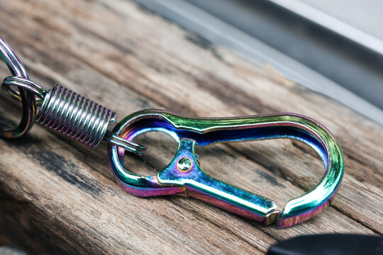 Iridescent metal snap hook with coiled spring resting on weathered wooden surface, detailed hardware showing vibrant color reflection and rugged outdoor utility design