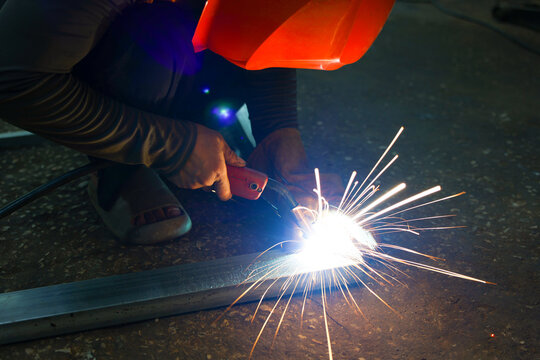 Skilled worker welding steel rail with bright sparks in dark workshop, wearing protective helmet and gloves during industrial metal fabrication work