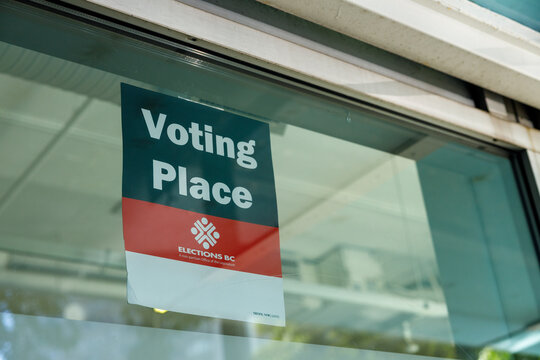 Elections BC Voting Place sign in dark green, red and white taped inside a glass window, marking a polling location, with ceiling reflections on the pane