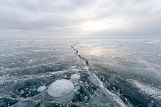 Frozen lake with a long crack and trapped air bubbles in clear ice under an overcast winter sky