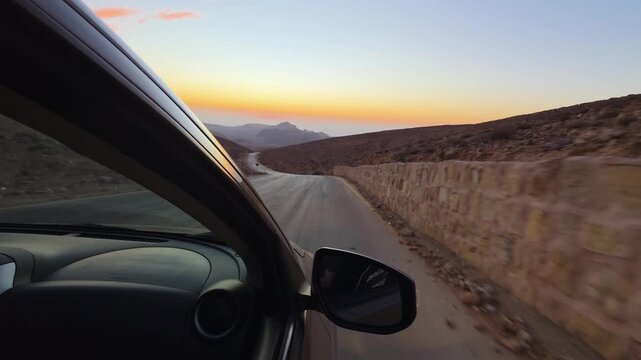 POV view from a moving car along a winding mountain road in Jordan at sunset, with arid hills, a warning sign and warm twilight glowing across the remote landscape.