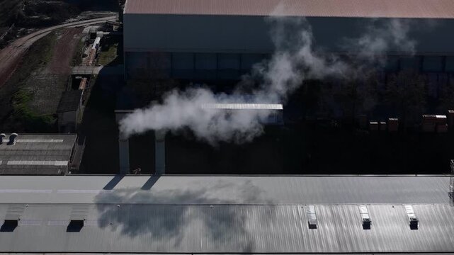 Industrial furnace emits dense white exhaust above a manufacturing building, illustrating industrial air pollution and fine particulate matter emissions, overhead drone view.